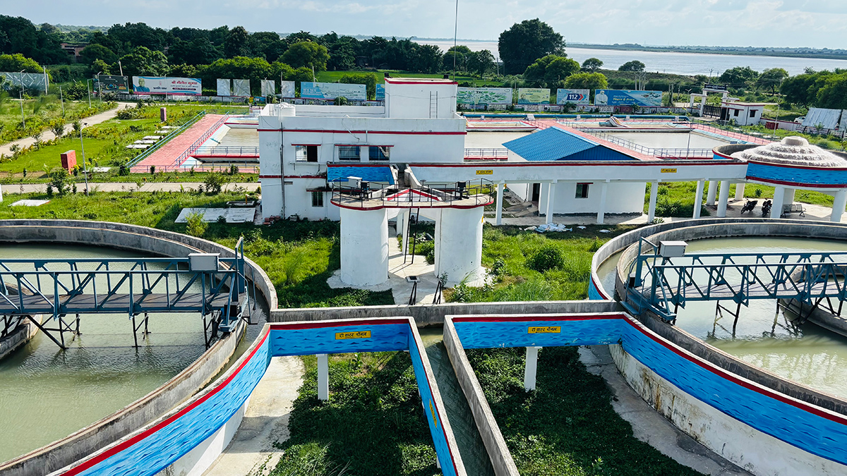 Two large concrete vats of water are seen from above at a water treatment plant. Various buildings and pathways are behind, and a body of water can be seen in the distance.