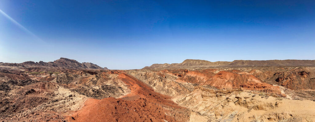 A panorama image showcases the hills, mountains, and valleys in the Turkana Rift Zone. Most of the landscape is a tan brown, and a red-orange streak of sediment cuts through the center of the image.