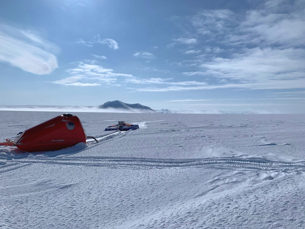 A red, triangular-shaped sled containing radar equipment is towed across an expansive ice sheet.