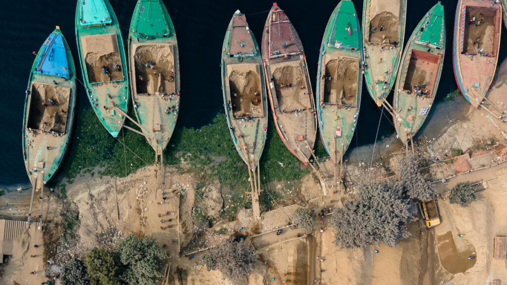Colorful boats filled sit side by side on a sandy bank, each with a line of trucks waiting to fill it with more extracted sand.