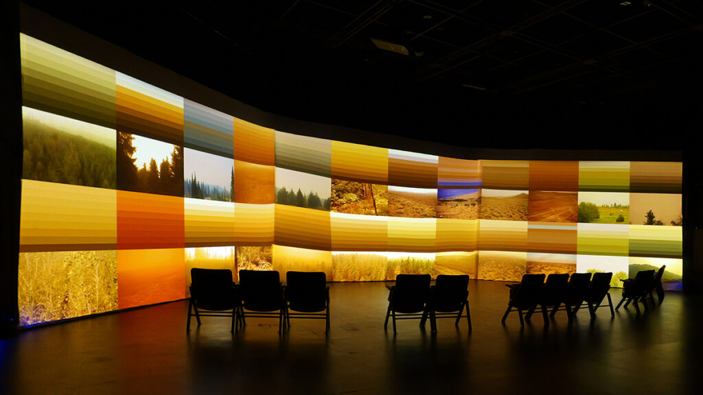 A row of 12 chairs, lined up in a dark room, is silhouetted against three screens showing orange-hued images. Some are just gradients of color, and others display landscapes.