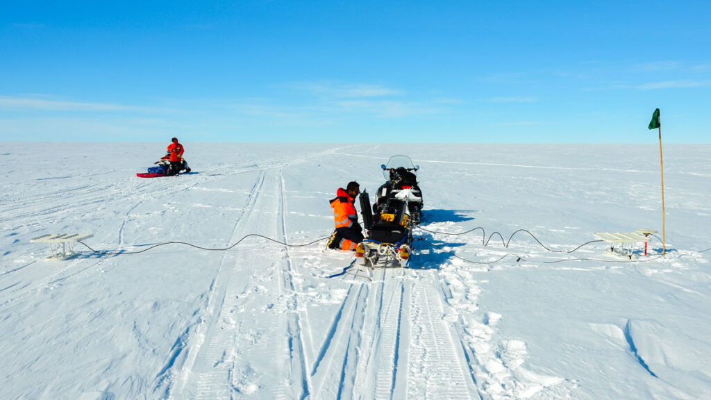 Amid a wide open expanse of snow-covered ice sheet under a blue sky, a researcher crouches beside scientific equipment set atop a sled behind a snowmobile.