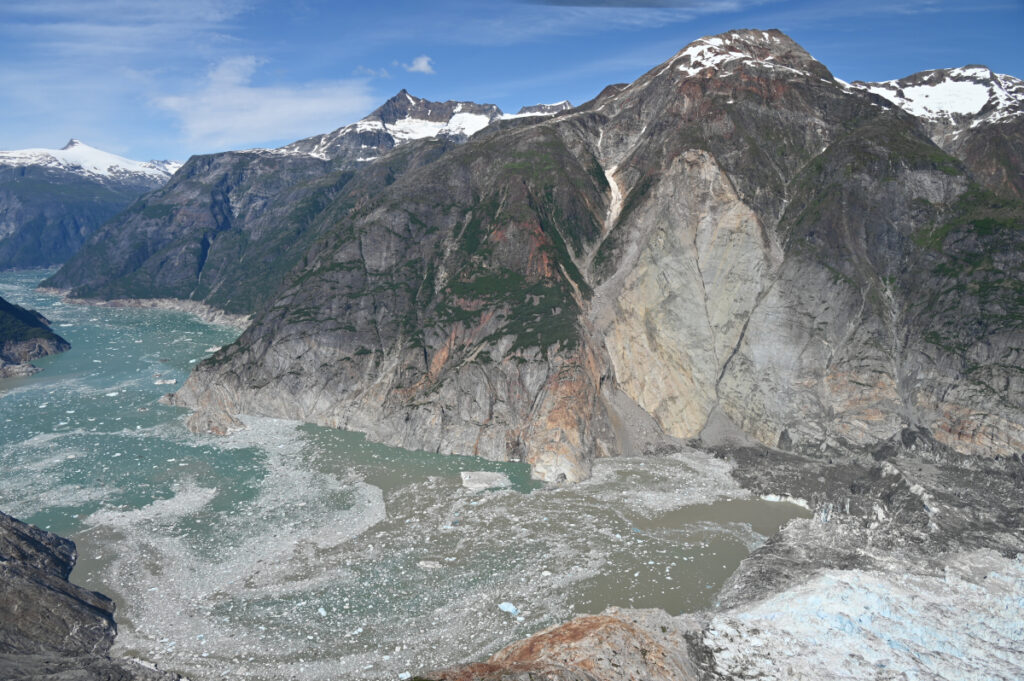 A photo shows a mountainside with a large wedge of lighter-colored rock, above a churning channel of water. The foot of a glacier can be seen at the lower edge of the image.