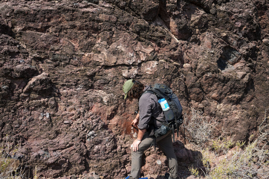 A scientist in desert gear points to a reddish patch of rock in a brown rock face.
