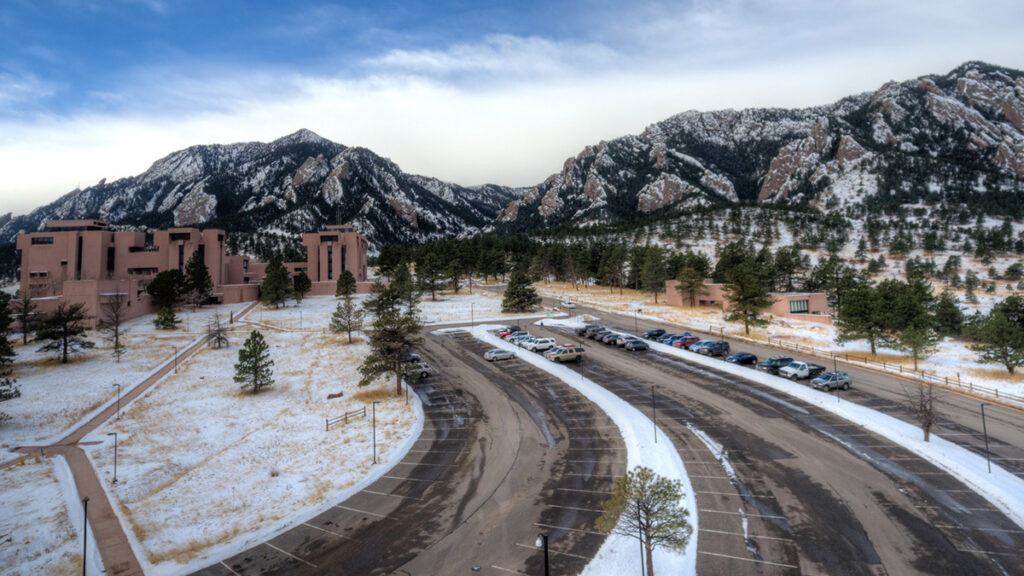The National Center for Atmospheric Research Building is seen on a snowy day, with the roads leading to it cleared, a few cars in the parking lot, and mountains just behind the building.