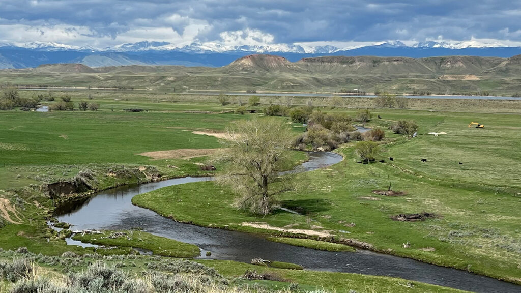 Panoramic view overlooking a stream meandering through green farm fields toward hills and mountains in the distance