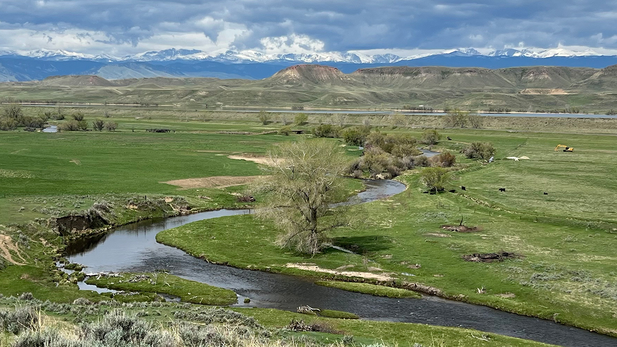 Panoramic view overlooking a stream meandering through green farm fields toward hills and mountains in the distance