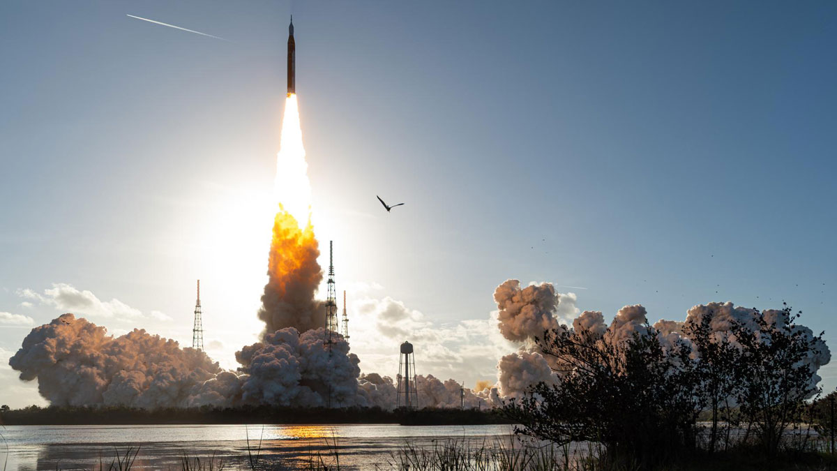 This image shows a rocket launching into a blue sky from its launchpad. A bright white and orange tail is emitted from the bottom of the rocket, transitioning into cloudlike billows of gas closer to the ground. A body of still water is in the midground, and grasses and shrubs are in the foreground.