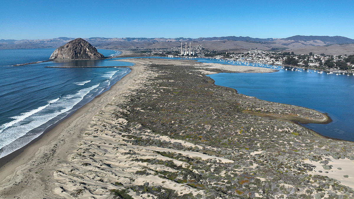 A sand bar on a beach is surrounded by water on both sides. A coastline with infrastructure is seen in the distance.
