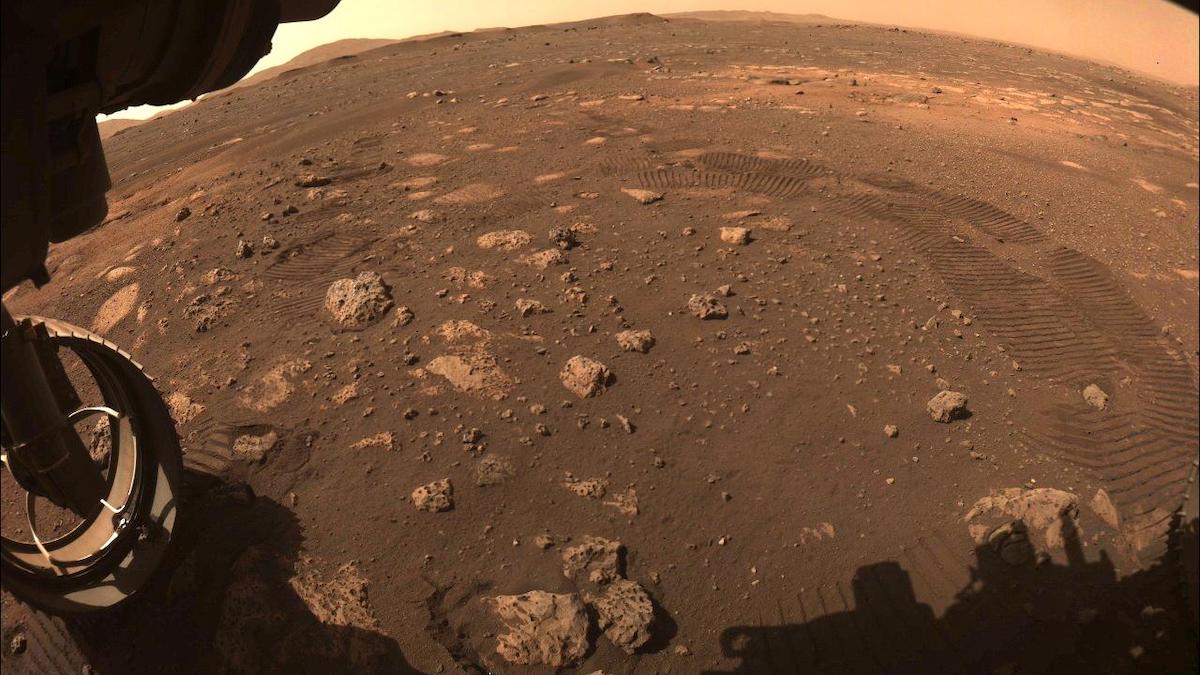 An image of Mars shows reddish-brown rocks and dust. The tracks of the Perseverance rover make an impression on the surface.