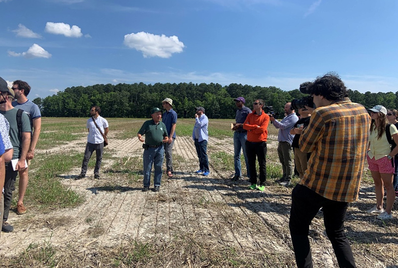 A group of roughly a dozen people stand in farm field listening to one of the farm owners talking. The field has patches of low green growth as well as large, bare patches where nothing is growing.