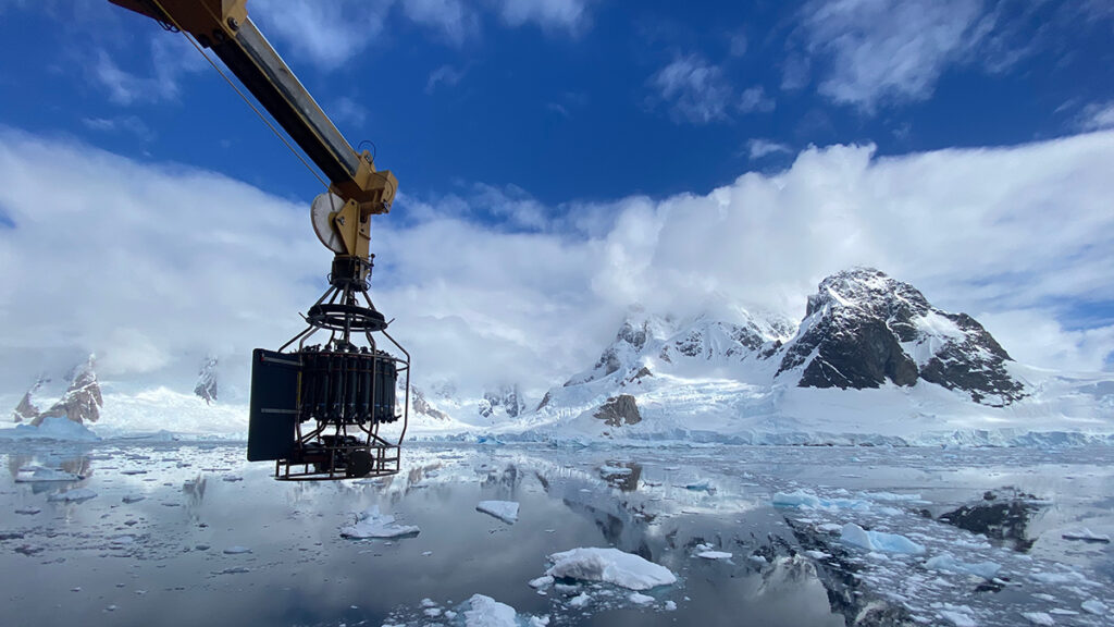 A metal arm with a cylindrical cage at the end of it extends off the side of a boat, over glassy water with chunks of ice in it. Snowy mountains and clouds are reflected in the water.