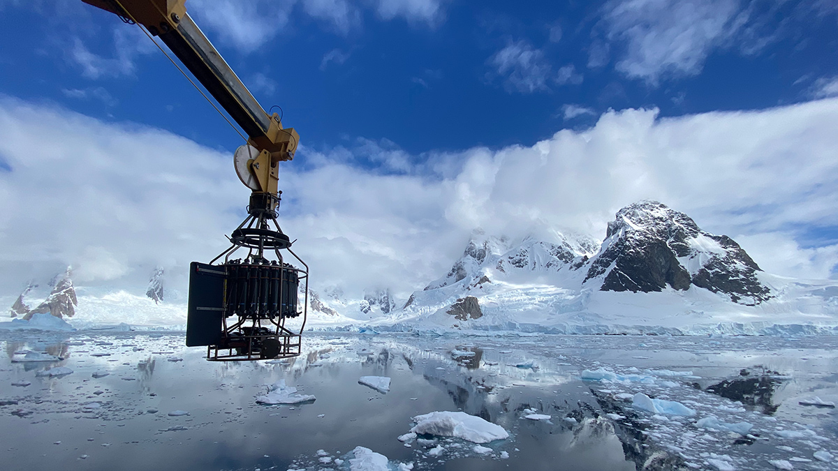 A metal arm with a cylindrical cage at the end of it extends off the side of a boat, over glassy water with chunks of ice in it. Snowy mountains and clouds are reflected in the water.