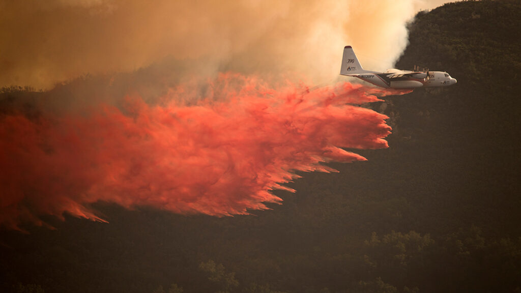 A plane flying over a cloud of smoke releases a load of red firefighting chemicals.