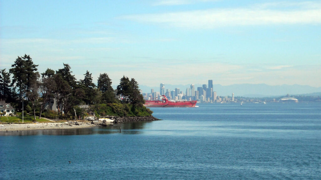 An island with evergreen trees and a sandy beach is seen in the foreground, while a mainland with tall buildings is in the background and a large red boat is in the midground.