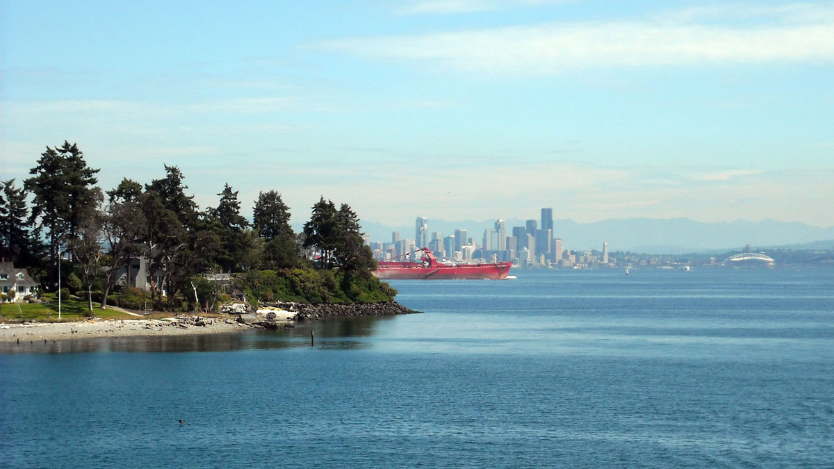 An island with evergreen trees and a sandy beach is seen in the foreground, while a mainland with tall buildings is in the background and a large red boat is in the midground.