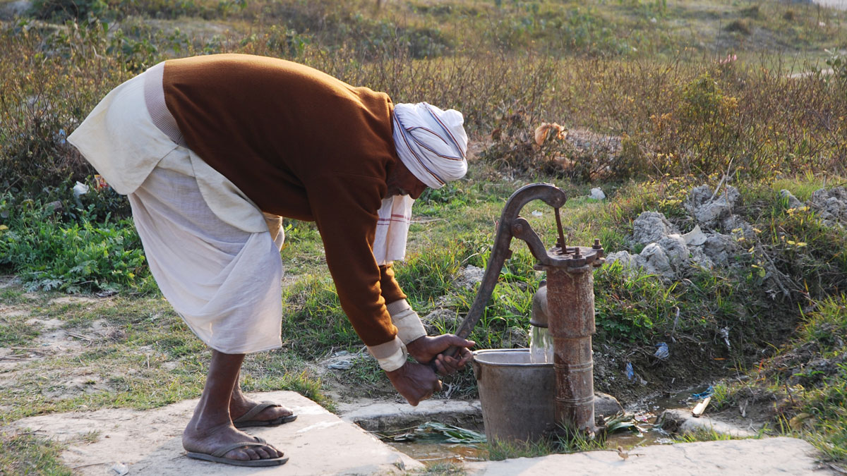 A man wearing flip-flops, loose pants, and a turban bends over to pump water into a metal bucket. There are shrubs and greenery behind him.