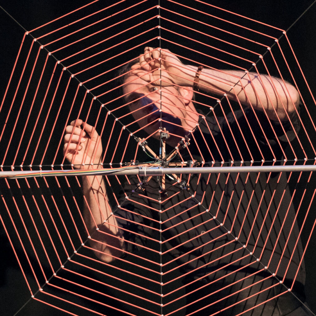 A man plays a large orange harp shaped like a spiderweb.
