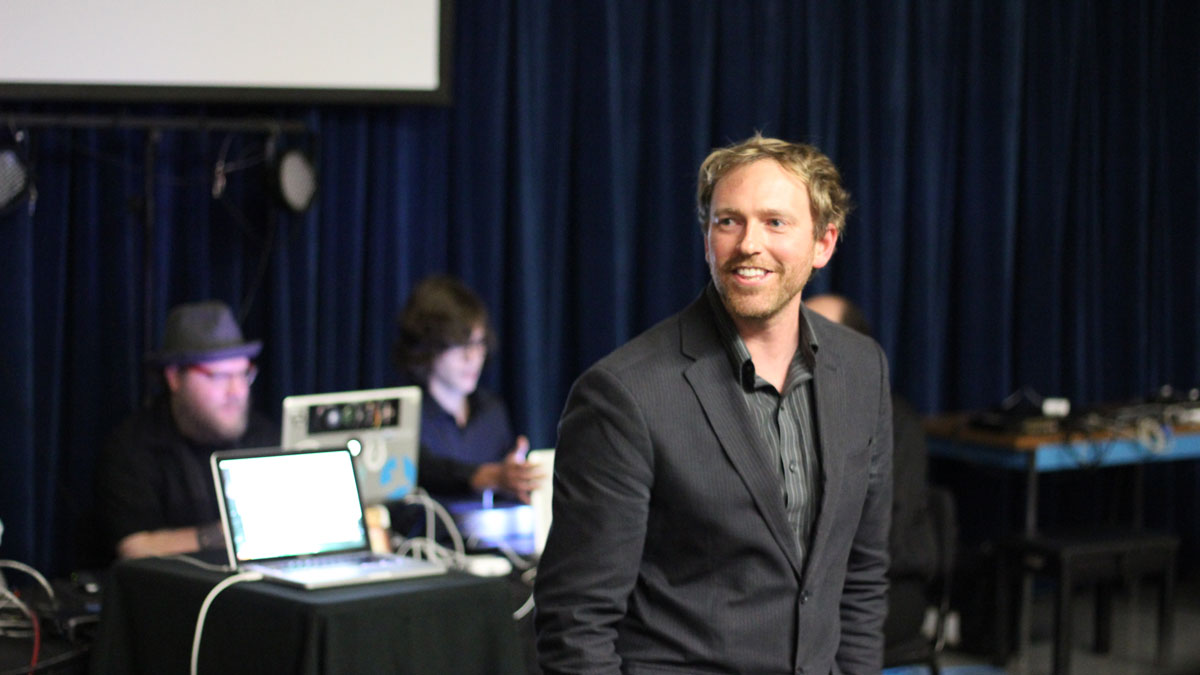 Chet Udell, dressed in a suit, stands in front of tables where people are using laptops.