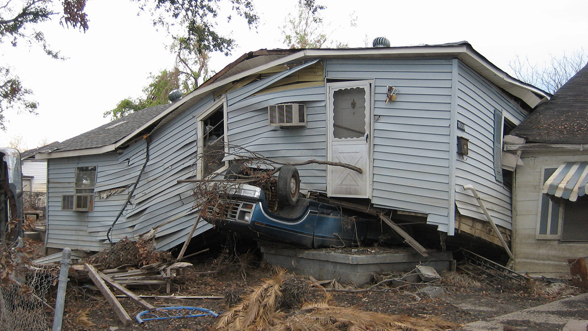 A displaced house sits on top of an upside-down car.