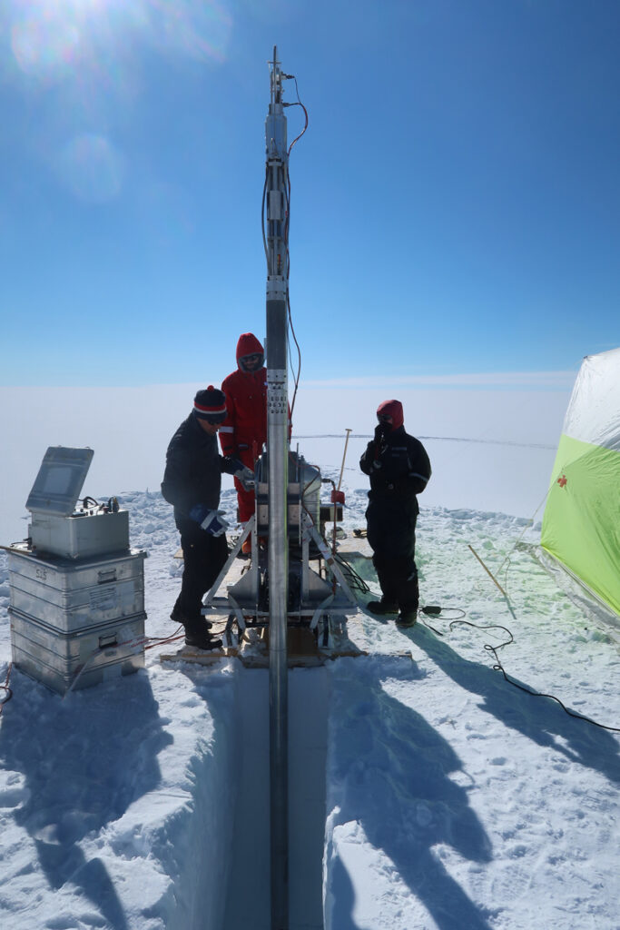 Three people gather around a long, tall metal object that is drilling a hole into the snow beneath them. A tent is visible behind them.