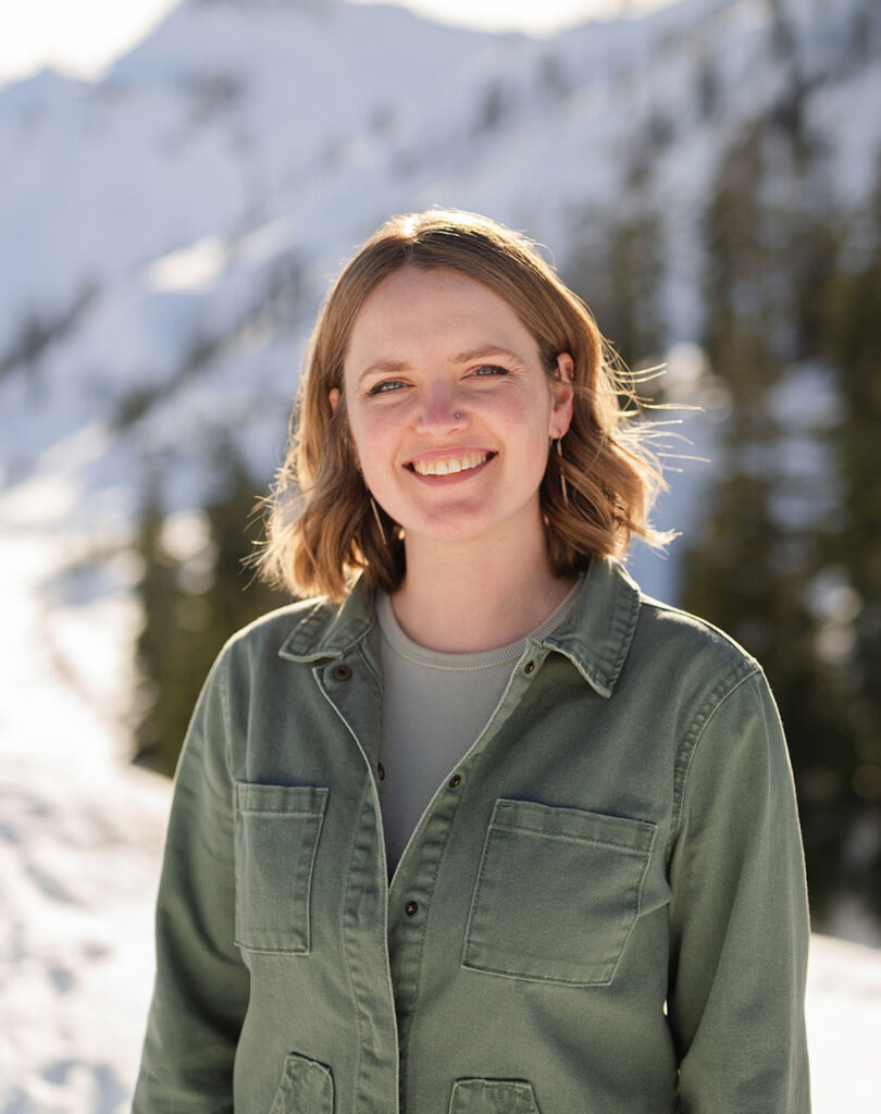 A woman wearing a green jacket stands and smiles in front of a snowy mountain landscape.