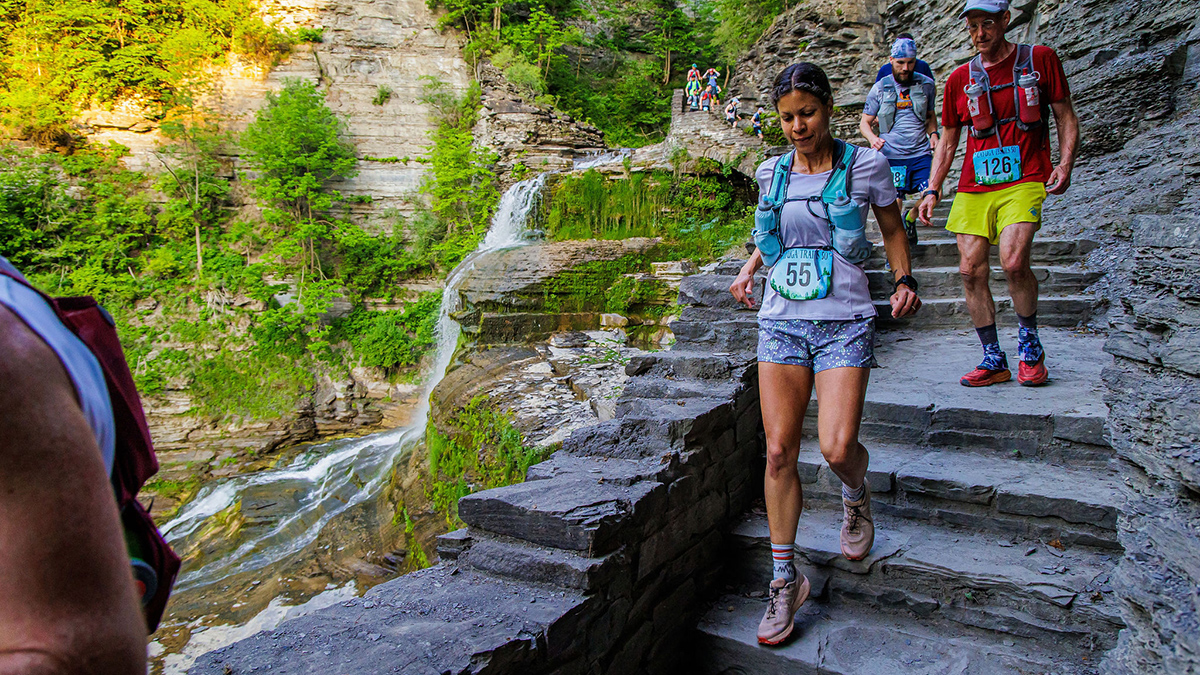 A group of people wearing hydration vests and race bibs run down a stone stairway alongside a cliff. A waterfall and greenery are visible in the background. Kuwanna Dyer-Pietras is in the center of the photo.