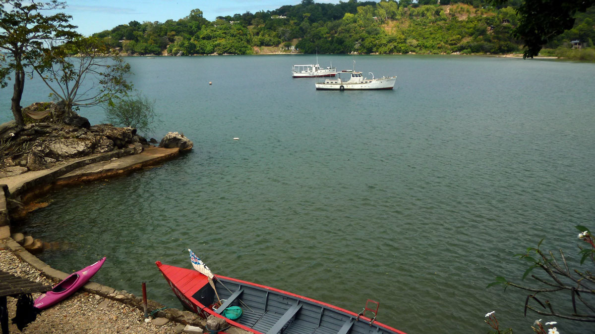 A small red boat is docked on the side of a lake, next to a wooden dock and a kayak. There are two motorized boats farther out on the lake and a hill covered in trees on the other side of the lake.