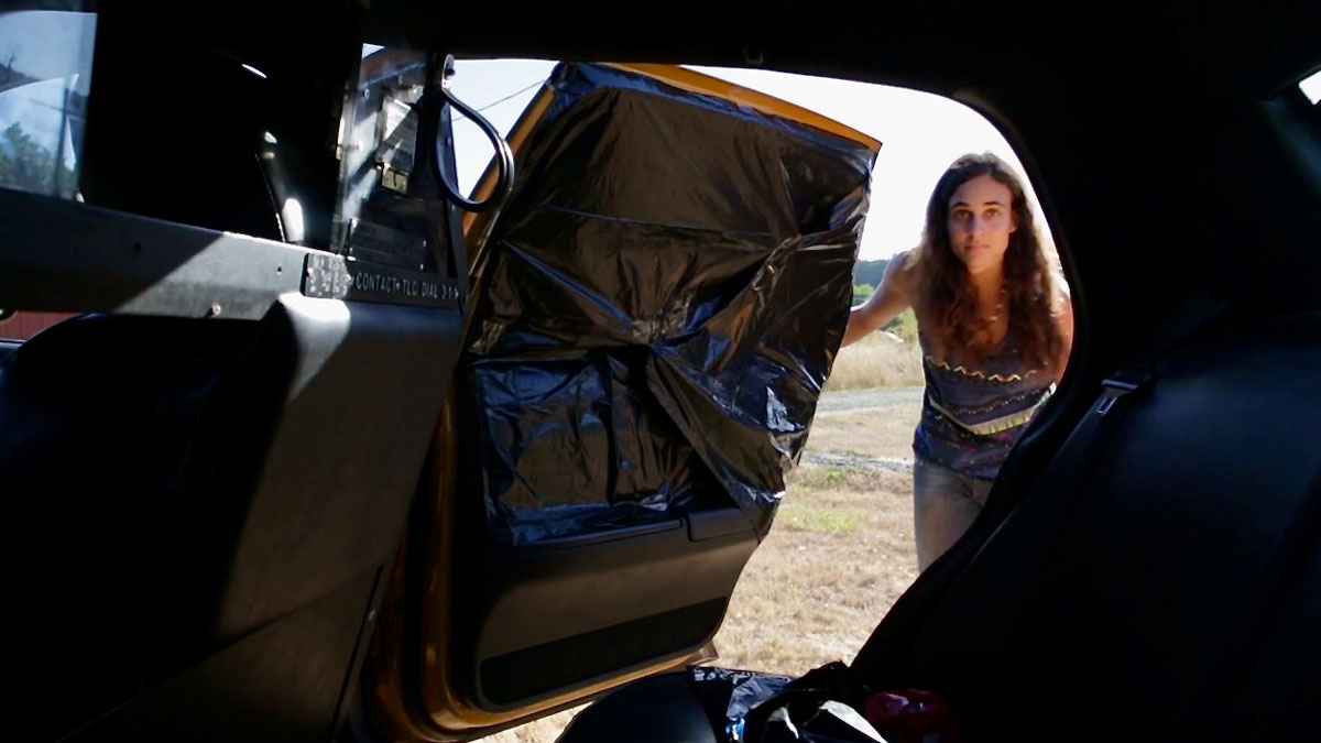 A young woman peeks into the back of a taxi cab. The photo, taken from inside the backseat, shows that the windows of the cab are covered in black trash bags.