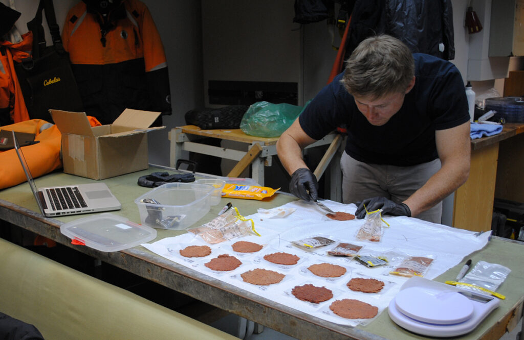 A person spreads orange, paste-like guano into thin layers at a lab bench.