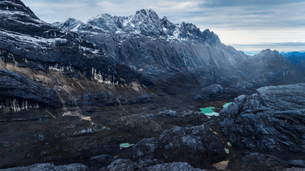 Rocky mountains, dusted in snow, fill most of the frame. The sky is cloudy, and three small spots of vibrant blue water are visible.