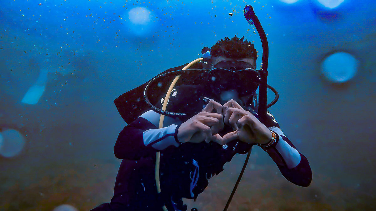 An underwater photo shows a young man in scuba gear making a heart symbol with his hands.