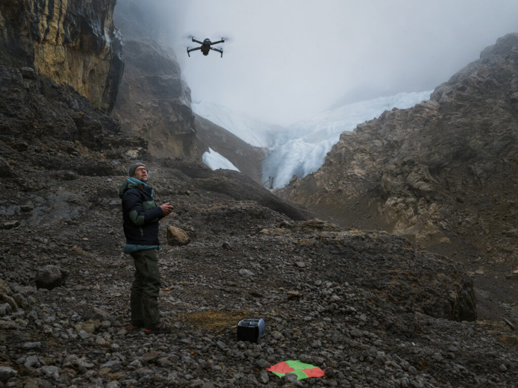 A man stands on a rocky mountain, looking up at a drone he is flying overhead. In the background, beyond the rocks, are a snowy mountain and a cloudy sky. An orange and green circle is on the ground next to the man.