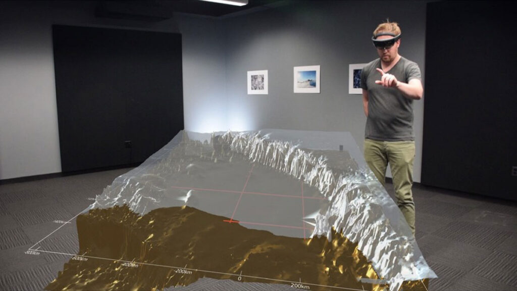 A person stands in an open room wearing virtual reality goggles, with their arm outstretched and index finger and thumb extended. In the foreground, a virtual reality depiction of Antarctic topography and ice are projected atop the photograph.