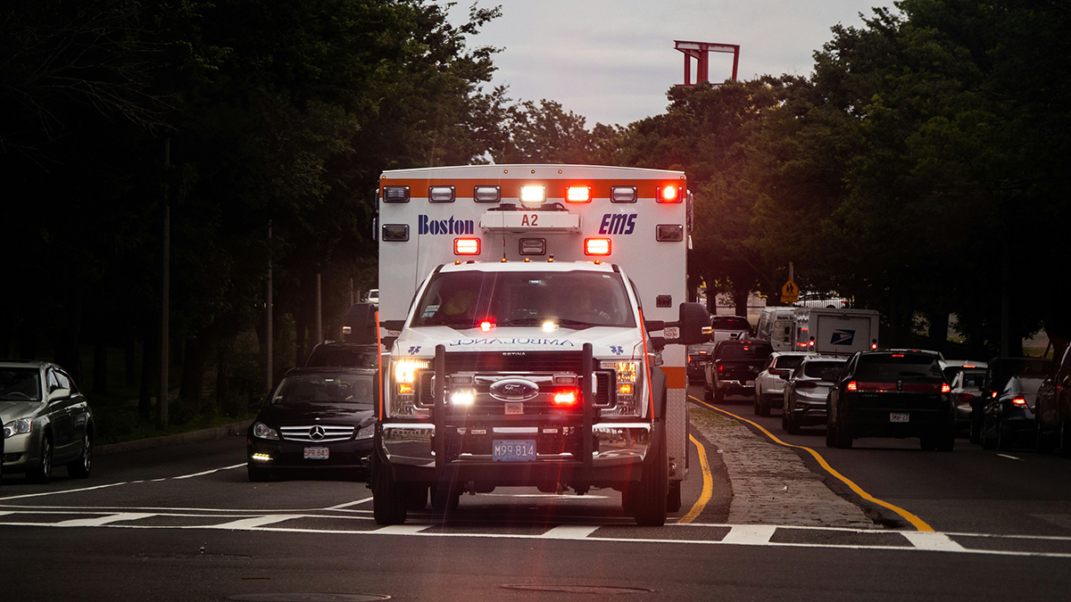 An ambulance with red lights shining drives down a road. The ambulance is labeled “Boston EMS.”