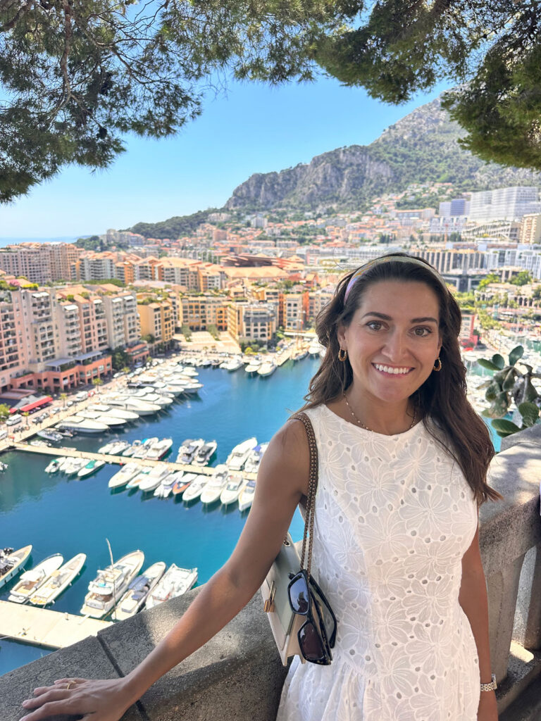A young woman in a white dress poses for a photo in front of a seaside town.