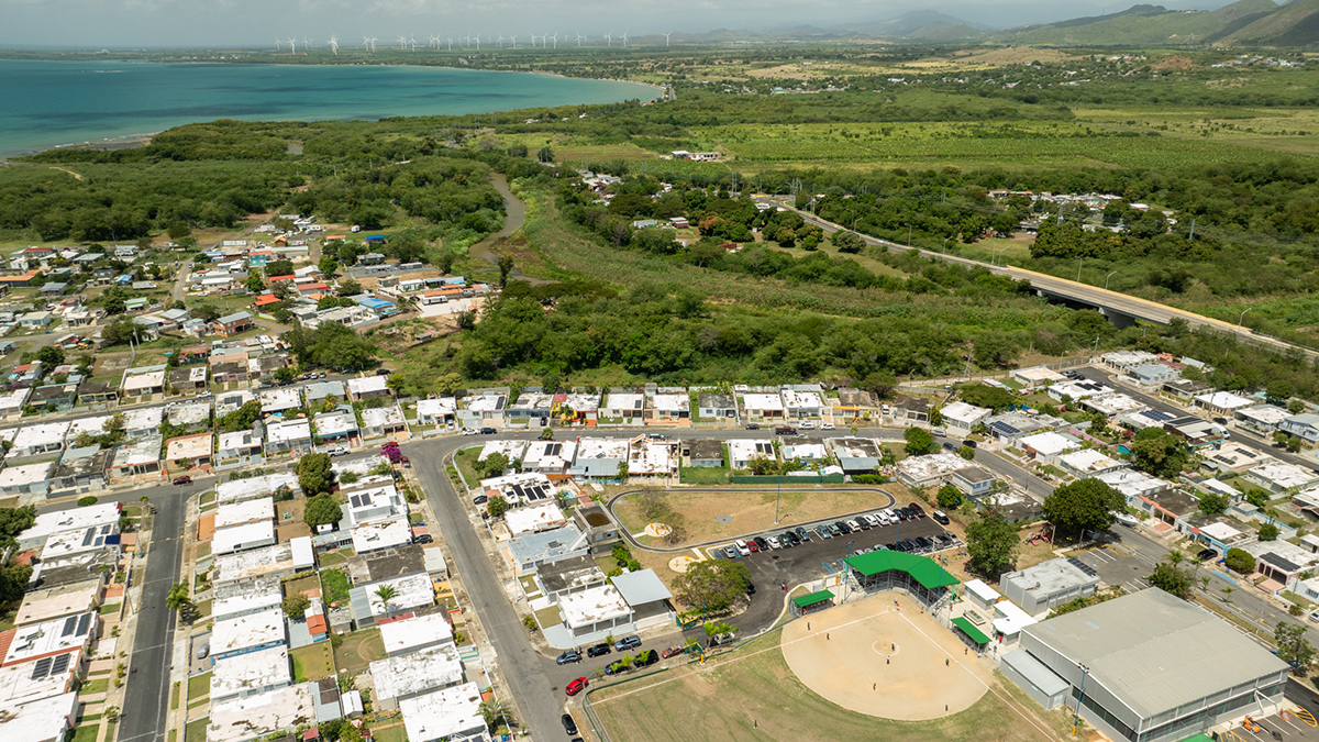 An aerial photo shows a city with low buildings in the foreground, a river and stretch of greenery in the midground, and blue ocean waters in the background.