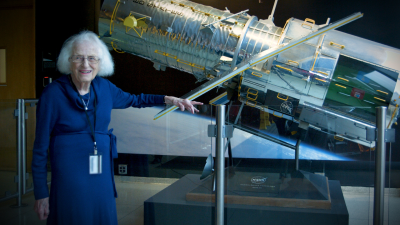 A woman in a blue dress stands in front of a scale model of a telescope with her arm resting on a glass half wall.