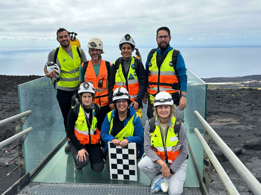 Seven people wearing yellow vests, most of whom are wearing hard hats, pose for a photo at an entrance to a lava tube.