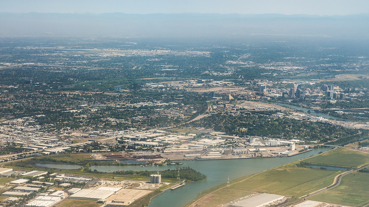 An aerial view of the city of Sacramento. High-rise buildings, a river, agricultural fields, and mountains can all be seen.