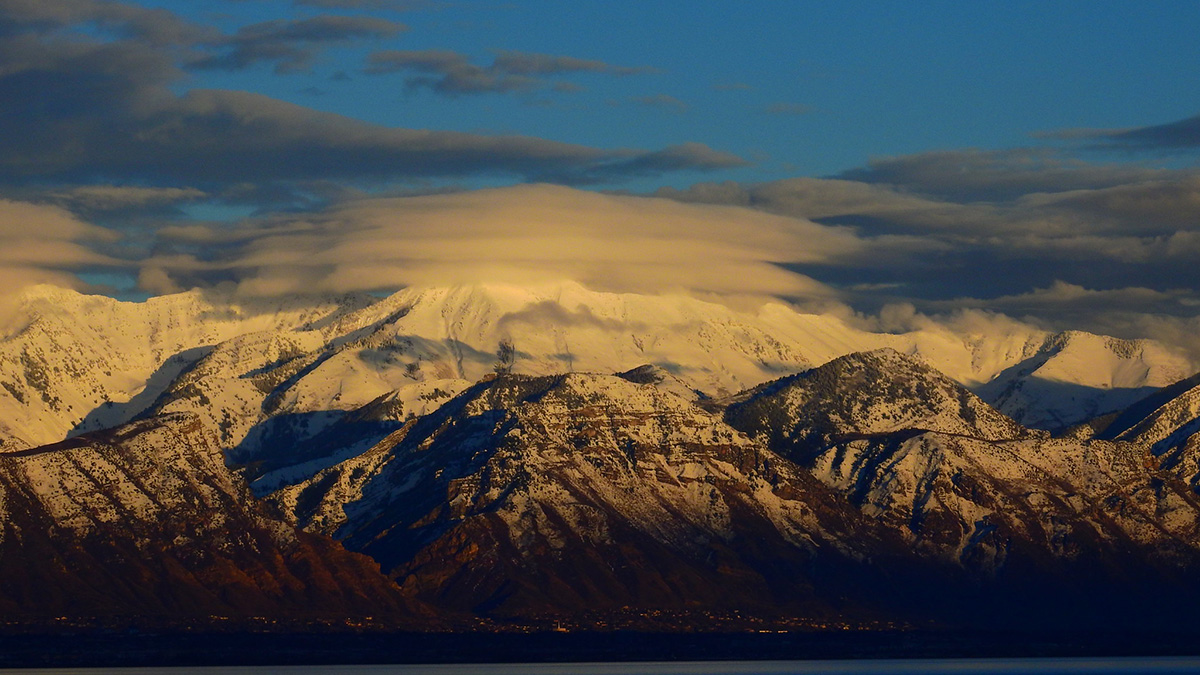 Clouds topped with snow, and with patchier snow lower down on the mountain, are beneath a long, low cloud.