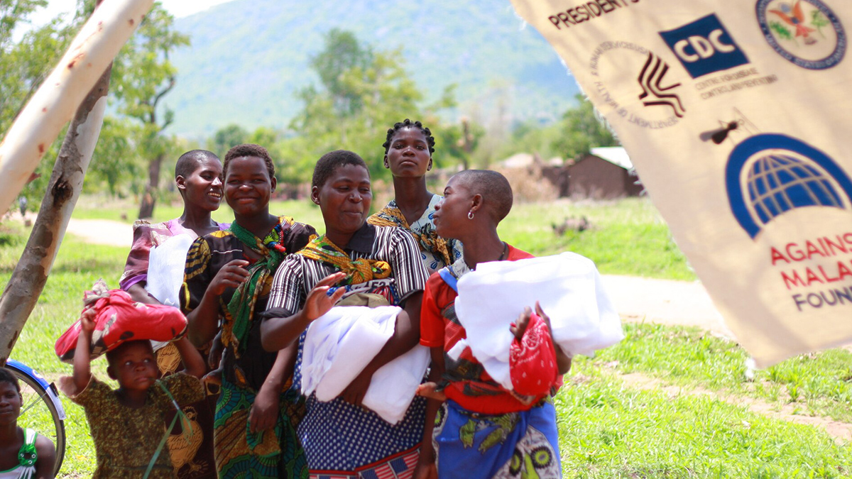 A group of five Malawian women and children hold antimalarial bed nets (folded white fabric) outside in front of a green landscape.