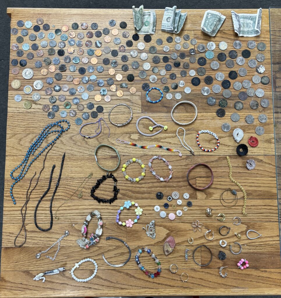 Dozens of coins and pieces of miscellaneous jewelry (icluding necklaces, bracelets, rings, and earrings), are laid out on a table. Also pictured are buttons and four dollar bills.
