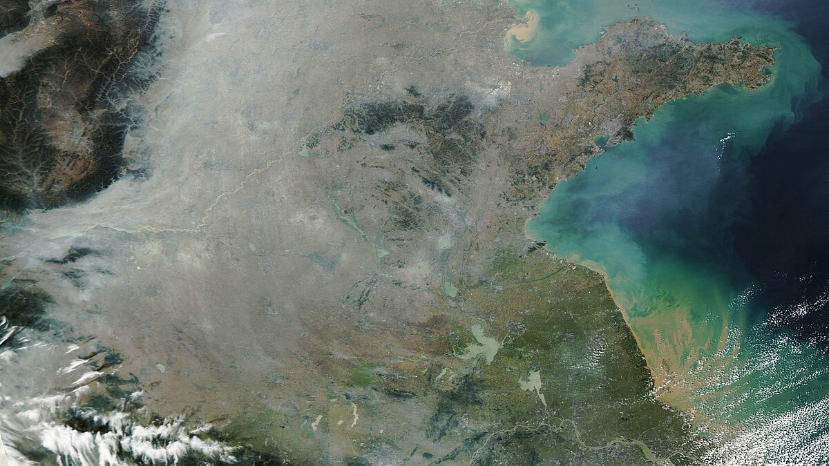 A satellite image shows a brown and green landmass with peninsulas and bays meeting a teal and blue body of water, with both partially overlain by a grayish haze.