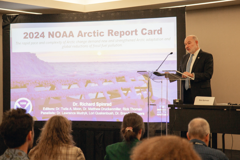 A scientist speaks at a podium in front of a projector screen that says "2024 NOAA Arctic Report Card."