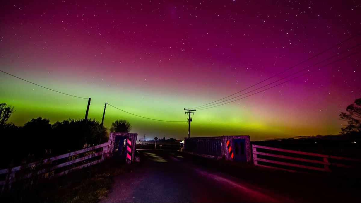 A starry night sky is visible through a rich magenta haze that smoothly blends into a bright yellow-green band near the horizon. In the foreground a road flanked by low railings and reflectors runs parallel to an electrical power line. Trees and a street sign appear in the distance.