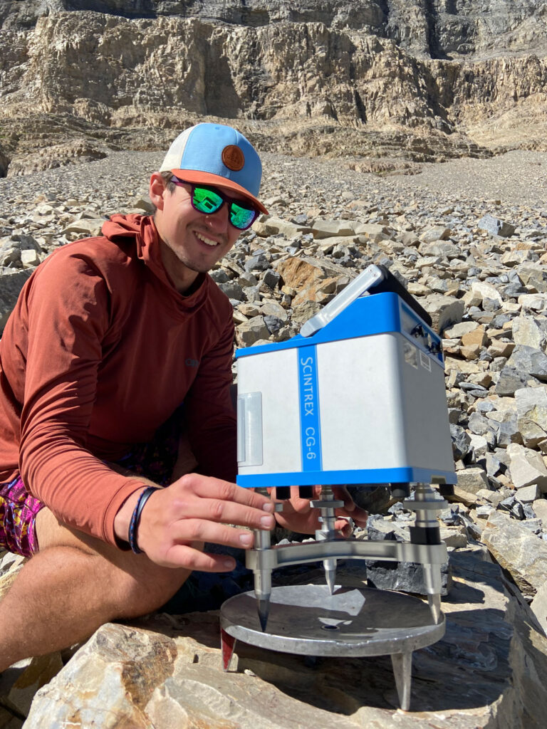 A man wearing an orange hoodie, a blue hat, and sunglasses holds a blue and gray metal contraption called a gravimeter, with rocks in the background.