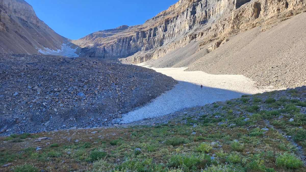 A rocky valley between two mountain ridges with a collection of snow at the bottom in partial Sun. A person is walking on the snow in the distance.
