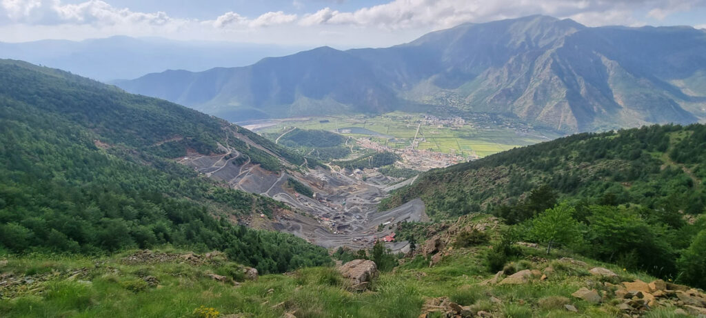 Panoramic view looking down a green hillslope toward a mine, where the vegetation is gone, revealing gray rock slopes. In the distance is a town situated in a flat valley expanse, and beyond the valley, high mountains rise.