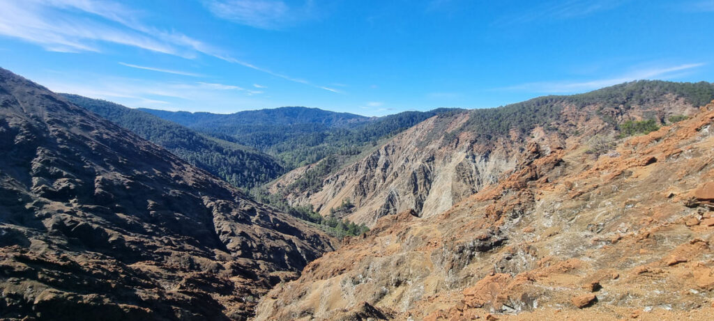 Orange- and brown-colored hillslopes dip toward each other, forming a narrow snaking valley between them. Tree-covered hills rise in the distance.